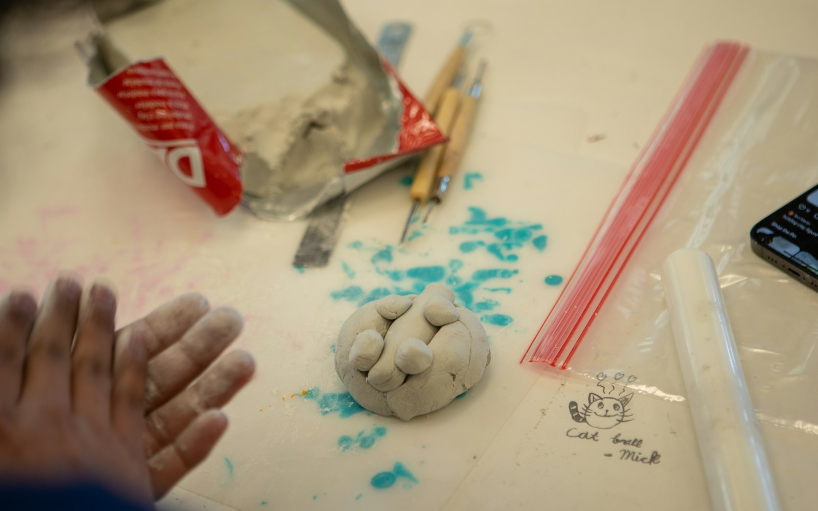 A small handmade air-dry clay doll drying on a work surface beside sculpting tools, acrylic paints, and a soft support cradle.