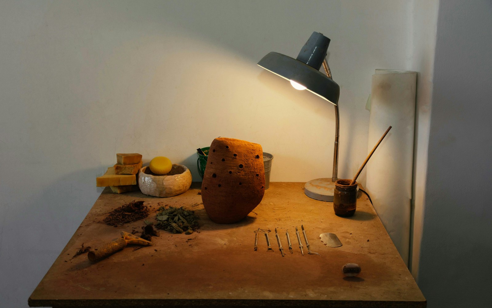 A worktable with unfinished air-dry clay doll faces, small sculpting tools, wire armature pieces, and painted sample parts.
