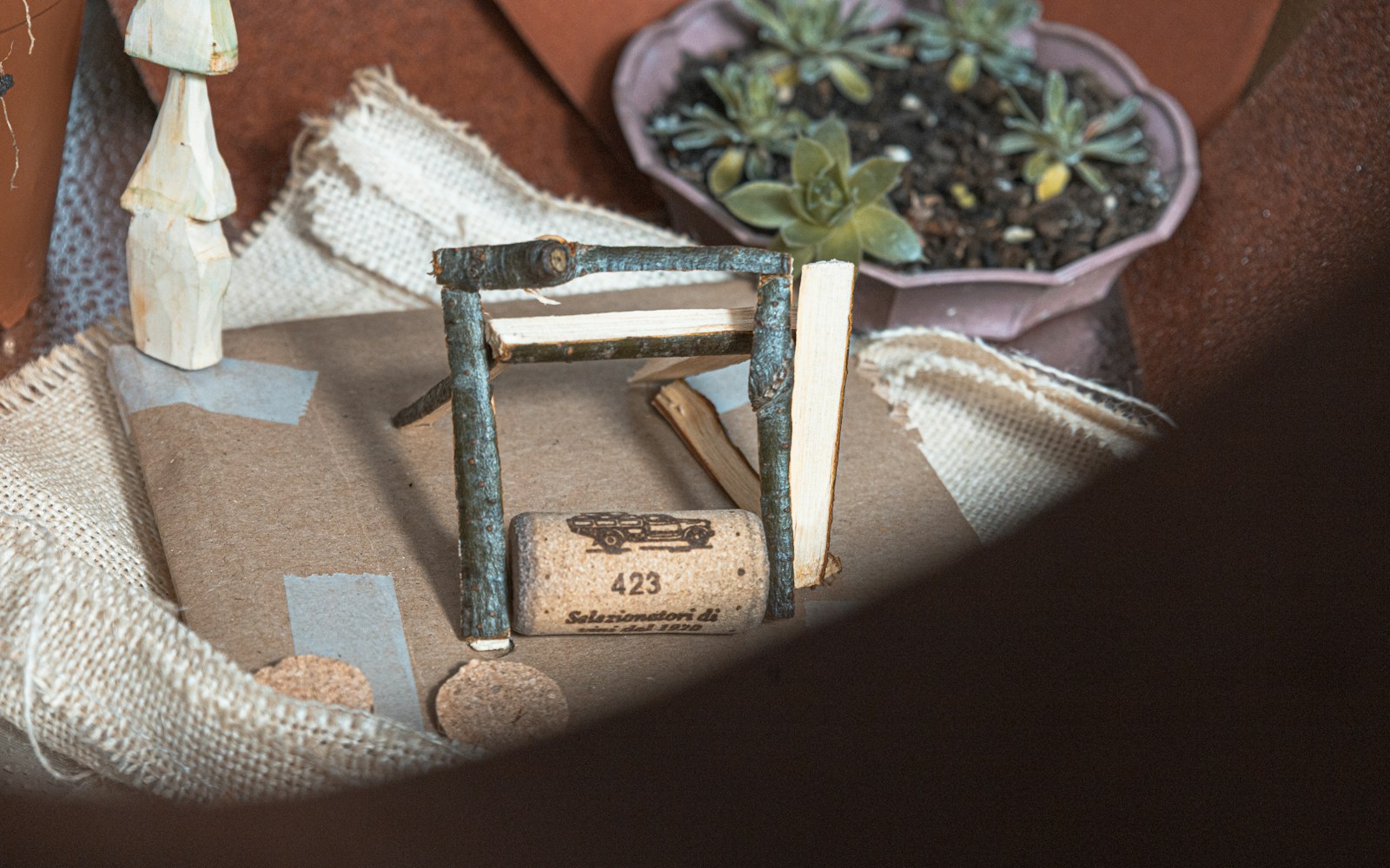 A miniature room box under construction with unpainted walls, paper templates, a ruler, and a small doll seated inside to check scale.