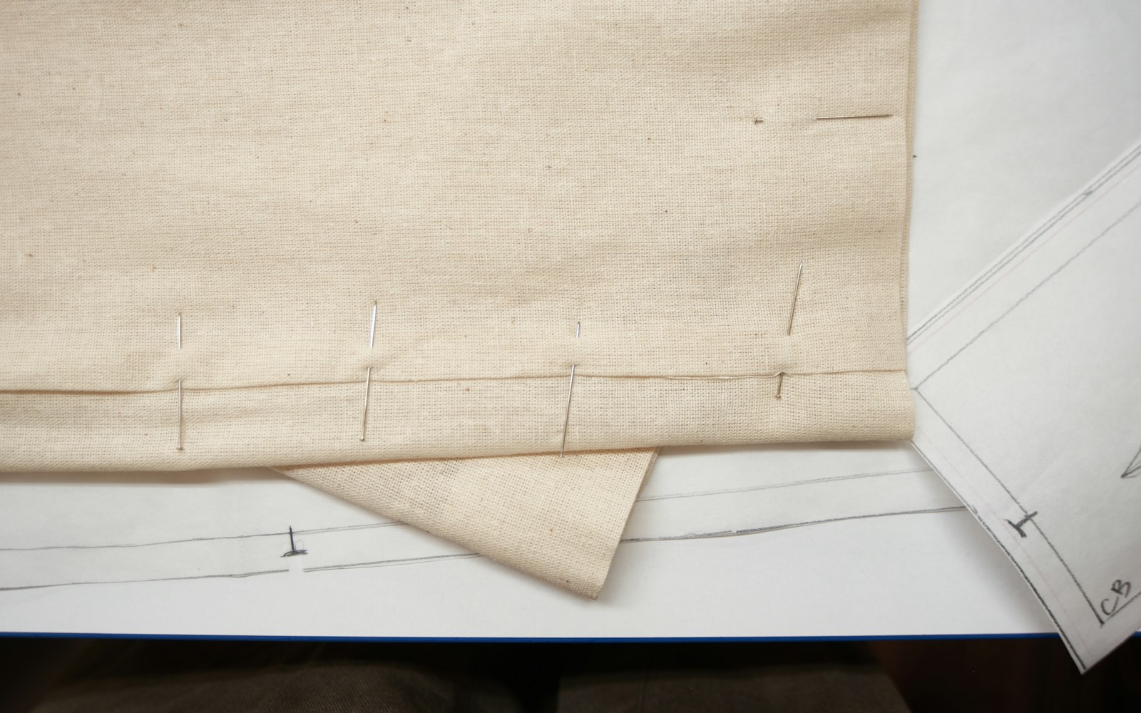 A worktable with a small dress form or art doll body, paper pattern pieces, unbleached muslin, fine sewing tools, and a partially constructed miniature gown.