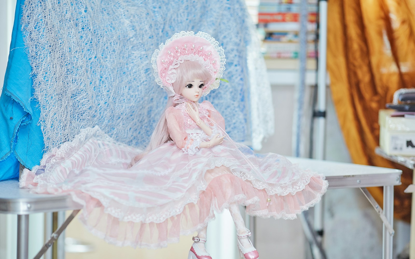 Close view of a maker fitting a tiny formal gown in pale fabric on an art doll, with pattern pieces, thread, and small trimming tools on a worktable.
