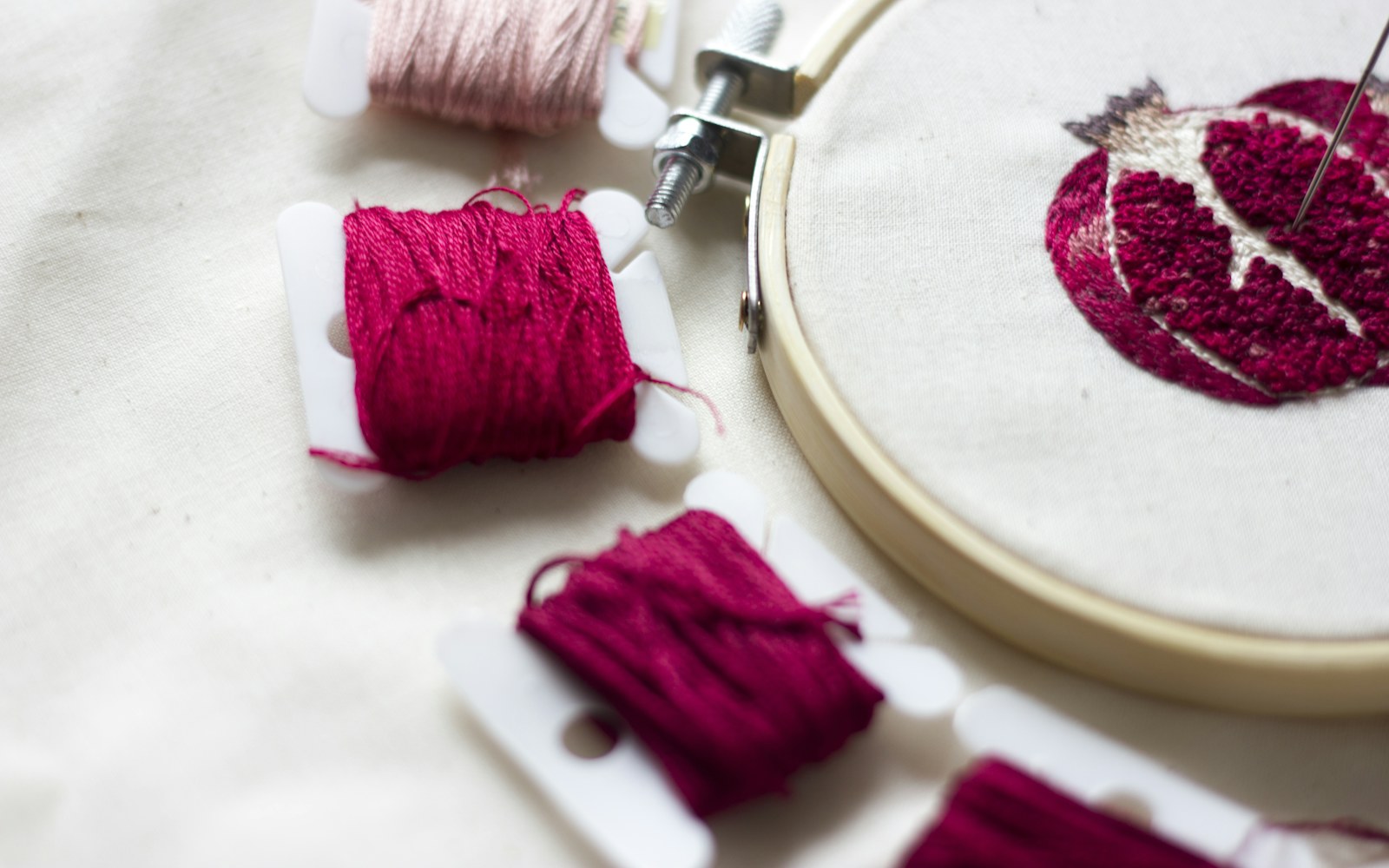 Close view of a handmade textile doll head in an embroidery hoop, with layered thread shading around the eyes and small beads placed as accents.