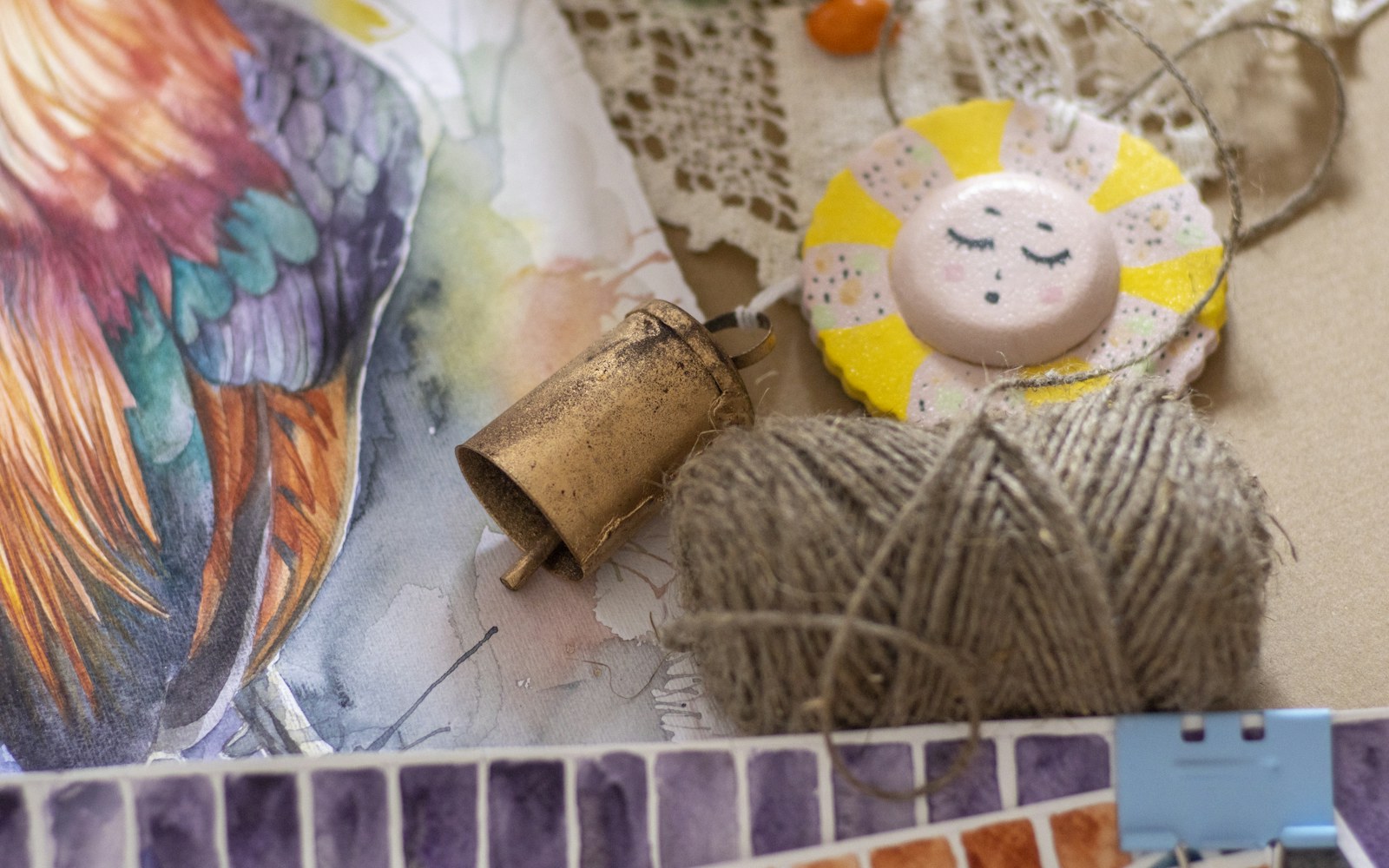 Small handmade doll face surrounded by clay, fabric, paper, beads, glass, and paint tools on a worktable