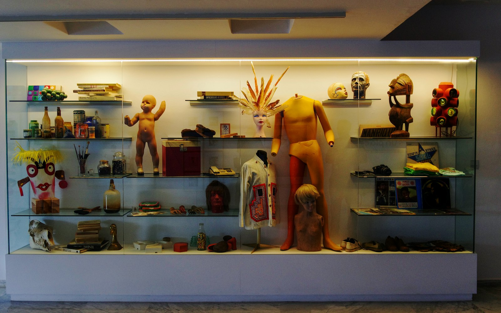 A softly lit textile doll displayed upright on a deep shelf beside a shadow box, with embroidery and dimensional details clearly visible.