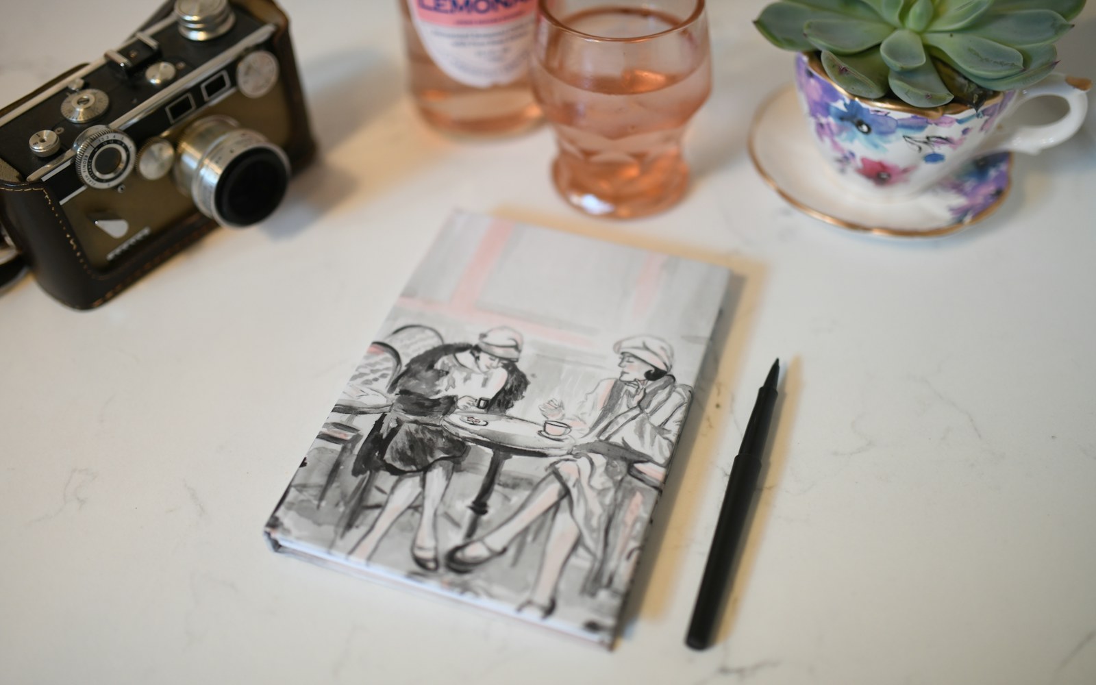 A handmade figure beside a notebook, measuring tape, photographs and archival storage materials on a studio table