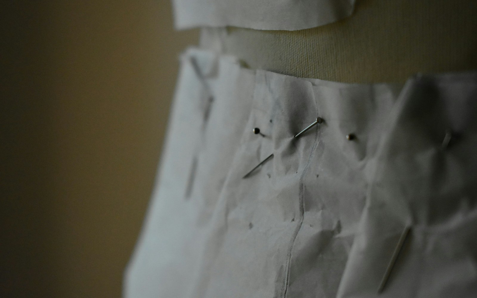 A handmade art doll on a worktable wearing a pale muslin gown mock-up, surrounded by small fabric swatches, thread, pins, and a historical dress reference.