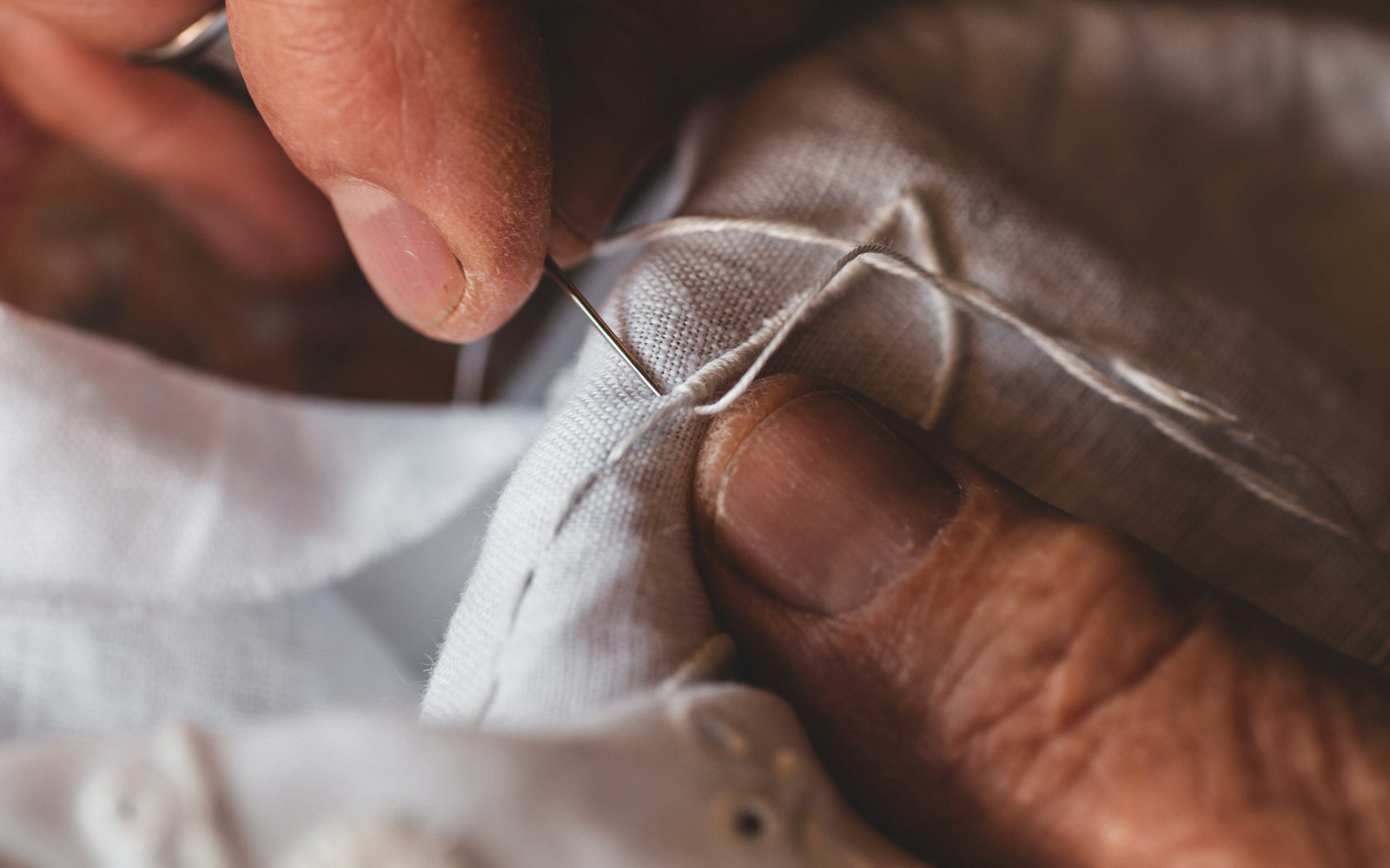 Close view of hands repairing a small embroidered cloth doll with needle, thread, fabric backing, and loose stuffing on a clean work surface.